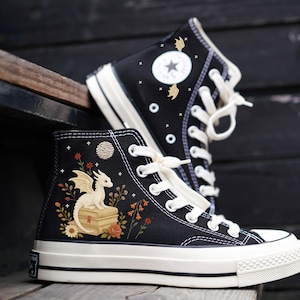 May include: Black high-top sneakers with white soles and laces. One shoe features a white dragon illustration with flowers and a moon. The other shoe has a star logo and bat illustrations. The sneakers are on a wooden surface.