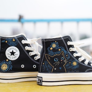 Puede incluir: Zapatillas altas negras con un diseño de gato y noche estrellada. Las zapatillas tienen una suela blanca, cordones blancos y el logo de Converse All Star. El diseño del gato es azul y amarillo.