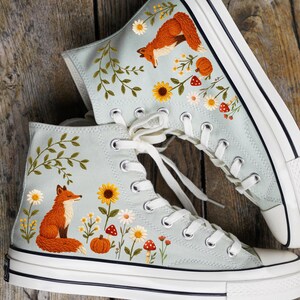 Op de afbeelding: Een paar lichtblauwe hoge sneakers met borduursels van een vos, zonnebloemen, madeliefjes, paddenstoelen en een pompoen. De schoenen hebben witte veters, een witte rubberen neus en een zwarte streep op de witte zool.