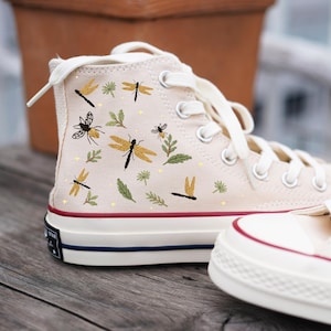 Könnte beinhalten: Cremefarbene High-Top-Sneaker mit Libellen- und Blätterstickerei. Die Sneaker haben weiße Schnürsenkel, einen rot-blauen Streifen auf der Sohle und ein schwarzes Sternlogo an der Seite. Das Design zeigt gelbe und schwarze Libellen und grüne Blätter.
