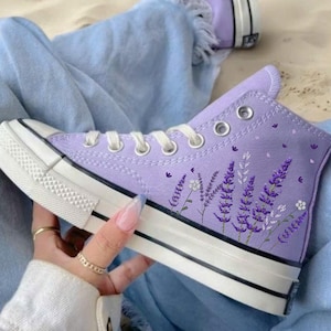 May include: Purple high-top canvas sneakers with a floral design featuring lavender flowers and white accents. The sneakers have white laces and a white rubber sole.