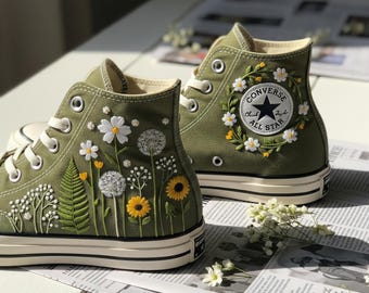 Puede incluir: Zapatillas altas verde oliva con diseños florales bordados. Las zapatillas tienen cordones blancos, suela negra y blanca, y un parche Converse All Star rodeado por una corona floral. Las flores bordadas incluyen margaritas, girasoles y dientes de león.