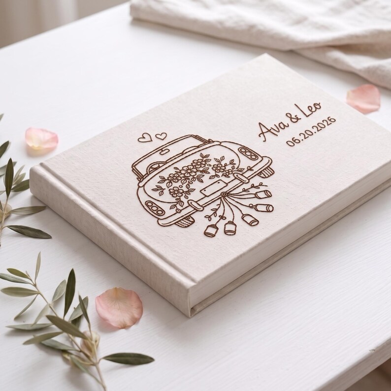 Puede incluir: Un &aacute;lbum de fotos rectangular cubierto de tela color hueso con un grabado l&aacute;ser marr&oacute;n oscuro de un coche antiguo decorado con flores y latas, con el texto Ava & Leo y la fecha 20.06.2026 grabados en la parte superior.