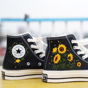 May include: Black high-top sneakers with white laces and soles. The sneakers feature a sunflower design on the sides, with yellow and brown flowers and green leaves. The Converse All Star logo is visible.