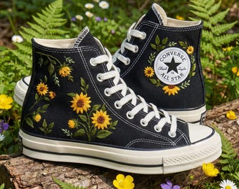 May include: Black high-top sneakers with embroidered yellow sunflowers and green leaves. The sneakers have white laces and a white rubber sole. The Converse All Star logo is on the side. The shoes are displayed on a log surrounded by flowers.