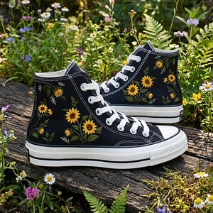 May include: Black high-top sneakers with white laces and soles. The sneakers are adorned with embroidered yellow sunflowers and green leaves. The shoes are set on a wooden surface surrounded by greenery and wildflowers.