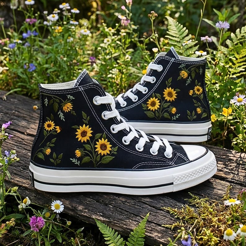 May include: Black high-top sneakers with white laces and soles. The sneakers are adorned with embroidered yellow sunflowers and green leaves. The shoes are set on a wooden surface surrounded by greenery and wildflowers.