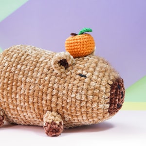 May include: A brown crocheted capybara plush toy with a small orange crocheted orange on top. The capybara is lying on a white surface.