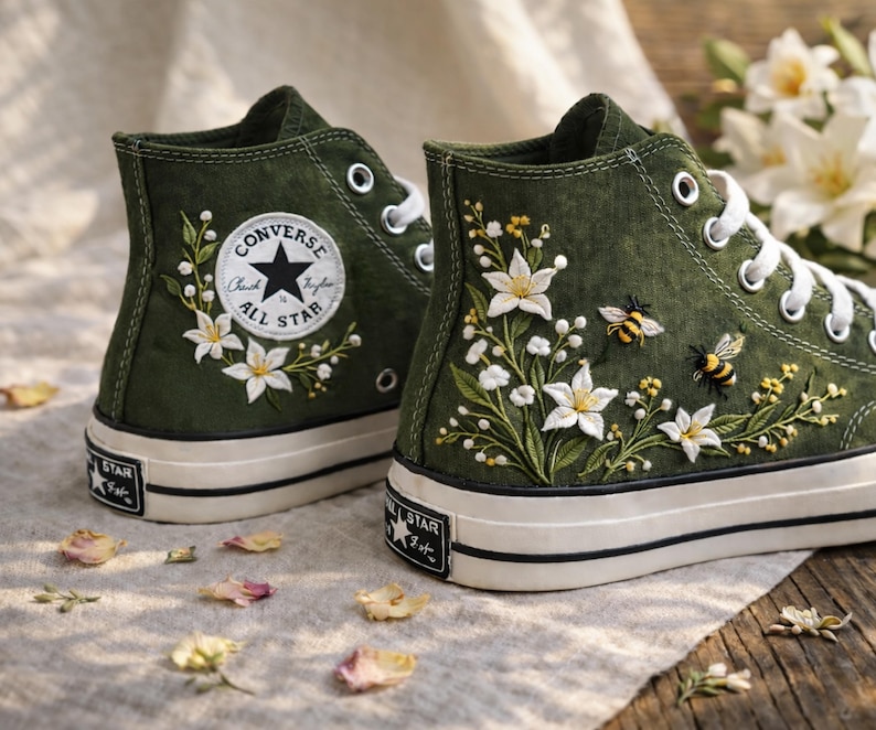 May include: Pair of high-top canvas sneakers in olive green, featuring white floral embroidery with bees. The sneakers have white laces, a white sole, and the Converse All Star logo on the side.