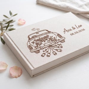 Puede incluir: Un &aacute;lbum de fotos rectangular de color blanco roto con un dise&ntilde;o grabado en marr&oacute;n de un coche antiguo decorado con flores y latas colgantes. El texto Ava & Leo y la fecha 20.06.2026 est&aacute;n grabados en marr&oacute;n en la parte derecha de la portada.