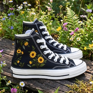 May include: A pair of black high-top sneakers with white laces and soles. The sneakers are decorated with yellow sunflower embroidery. The shoes are set on a wooden surface with a background of flowers and greenery.