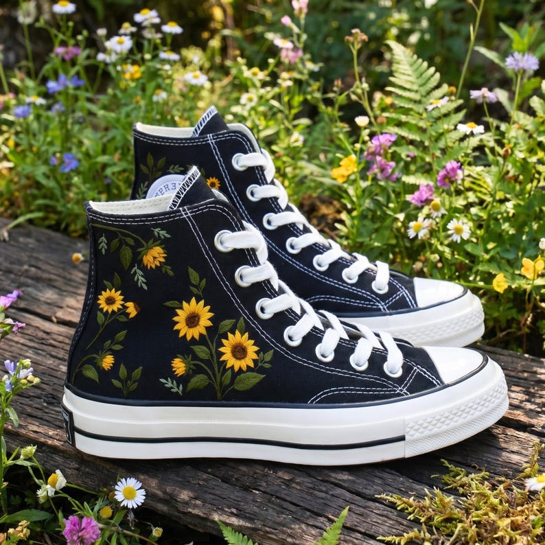 May include: A pair of black high-top sneakers with white laces and soles. The sneakers are decorated with yellow sunflower embroidery. The shoes are set on a wooden surface with a background of flowers and greenery.