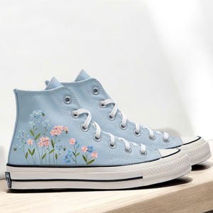 May include: Light blue high-top sneakers with floral embroidery. The sneakers feature white laces, a white rubber sole, and a black stripe. The embroidered flowers are pink, blue, and green.