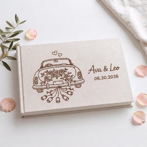 May include: A rectangular, light beige fabric-covered guest book featuring a dark brown engraved illustration of a vintage car decorated with flowers, two hearts above it, and tin cans trailing behind. The text reads Ava & Leo and 06.20.2026.