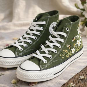 May include: A pair of high-top canvas sneakers in olive green. The sneakers feature white laces, a white rubber toe cap, and a white sole. One shoe has a floral and bee embroidery design. The Converse logo is visible on the side.