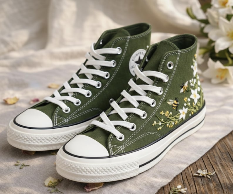 May include: A pair of high-top canvas sneakers in olive green. The sneakers feature white laces, a white rubber toe cap, and a white sole. One shoe has a floral and bee embroidery design. The Converse logo is visible on the side.