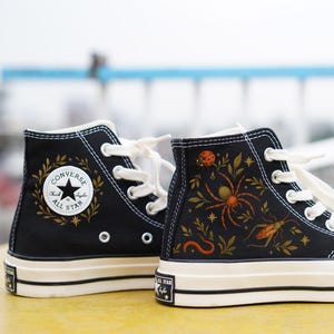 Könnte beinhalten: Schwarze High-Top-Sneakers mit weißen Schnürsenkeln und Sohlen. Ein Schuh hat einen goldenen Kranz mit dem Text "Converse All Star". Der andere Schuh hat eine detaillierte Illustration einer Spinne, eines Insekts und Blättern.