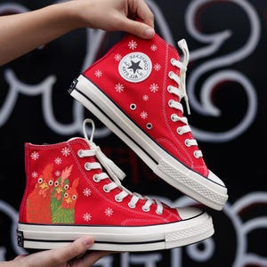 Op de afbeelding: Rode high-top sneakers met witte veters en zolen. De sneakers hebben een sneeuwvlokpatroon en een hanenontwerp. Het Converse-logo is zichtbaar aan de zijkant. De schoenen worden door handen omhoog gehouden.