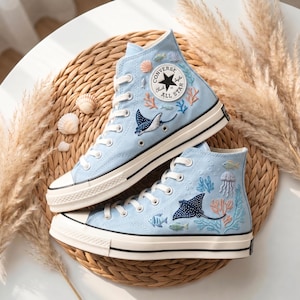 Könnte beinhalten: Hellblaue High-Top-Sneaker mit gestickten Meeresmotiven, darunter Mantarochen, Fische und Korallen. Die Sneaker haben weiße Schnürsenkel, einen schwarzen Streifen auf der Sohle und einen klassischen Converse All Star-Aufnäher.