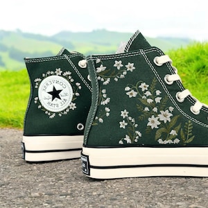 May include: A pair of dark green high-top canvas trainers with white floral embroidery and a black and white Converse All Star logo. The trainers have a white rubber sole.