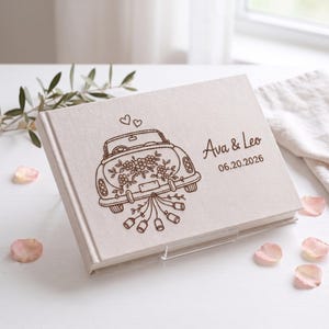 Puede incluir: Un libro de visitas rectangular cubierto de tela color hueso con un grabado l&aacute;ser marr&oacute;n oscuro de un coche descapotable vintage decorado con flores y latas colgantes, con el texto Ava & Leo y la fecha 06.20.2026.