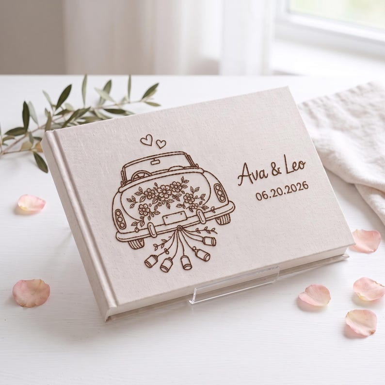 Puede incluir: Un libro de visitas rectangular cubierto de tela color hueso con un grabado l&aacute;ser marr&oacute;n oscuro de un coche descapotable vintage decorado con flores y latas colgantes, con el texto Ava & Leo y la fecha 06.20.2026.