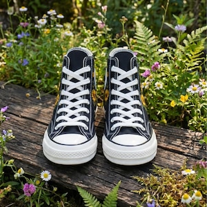 May include: Pair of black high-top sneakers with white laces and soles. The sneakers feature a yellow sunflower design on the sides. The shoes are placed on a wooden surface surrounded by flowers and greenery.