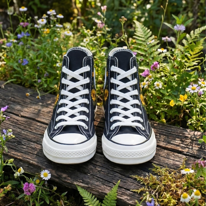 May include: Pair of black high-top sneakers with white laces and soles. The sneakers feature a yellow sunflower design on the sides. The shoes are placed on a wooden surface surrounded by flowers and greenery.