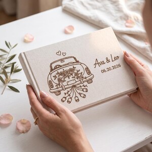 Puede incluir: Un &aacute;lbum de fotos rectangular cubierto de tela color hueso con una ilustraci&oacute;n grabada con l&aacute;ser en marr&oacute;n oscuro de un coche antiguo decorado con flores y latas, con el texto Ava & Leo y la fecha 06.20.2026 grabados en la portada.