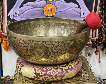 May include: A large, ornate brass singing bowl with intricate floral and geometric patterns. The bowl rests on a purple and gold cushion, with a wooden mallet and red felt striker inside. The background includes prayer beads and a spiritual artwork.