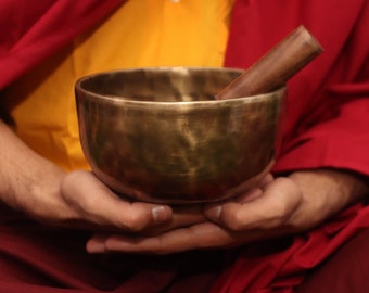 May include: A person holding a large, antique-looking, brass singing bowl with a wooden striker. The bowl is resting in their hands, and the striker is resting on the rim of the bowl.