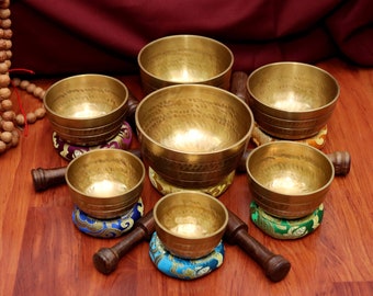 May include: A set of seven singing bowls, each with a unique design and color, arranged in a circle on a wooden surface. The bowls are made of metal and have a smooth, polished finish. Each bowl is accompanied by a wooden striker.