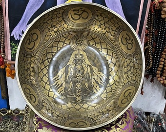 May include: A large, round, brass singing bowl with intricate gold designs. The bowl features a central image of a deity in a seated meditation pose, surrounded by Om symbols. The bowl is a traditional Tibetan singing bowl used for meditation and sound healing.