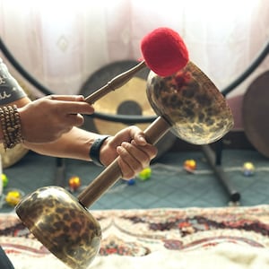 May include: A person plays a set of two singing bowls with a wooden mallet topped with a red felt striker. The bowls are decorated with a hammered finish and have a brown and gold colour.