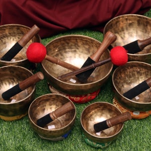 Może przedstawiać: Zestaw siedmiu starożytnych śpiewających misek z drewnianymi młotkami i czerwonymi filcowymi uderzaczami. Miski są ułożone w półkole na zielonej trawiastej powierzchni.