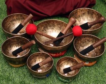 May include: A set of seven antique-looking singing bowls with wooden mallets and red felt strikers. The bowls are arranged in a semi-circle on a green grassy surface.