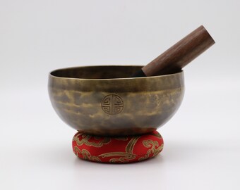 May include: A brass singing bowl with a wooden striker and a red cushion with a gold cloud design. The bowl has a decorative design on the side.