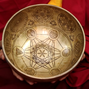May include: A gold singing bowl with intricate geometric designs etched into the surface. The bowl is held in the hands of a person wearing a red robe.