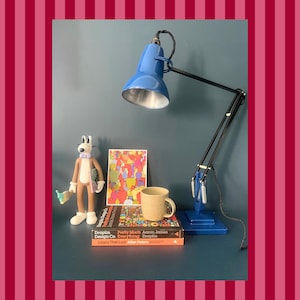Op de afbeelding: Een blauwe bureaulamp met een zilveren binnenkant, naast een cartoonfiguur, een kleurrijke abstracte kunstdruk, een gespikkelde mok en twee boeken. De arm van de lamp is zwart en de basis is blauw. De achtergrond is donkerblauwgroen.