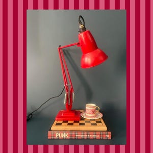 May include: A red desk lamp with a flexible arm and a round shade. The lamp sits on a red base, with a small teacup and saucer on a wooden surface. The word "PUNK" is visible on a book below.