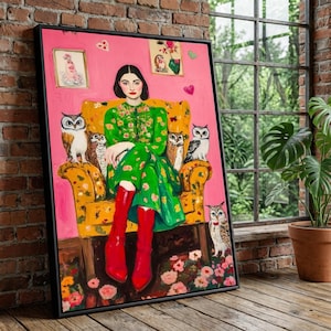 May include: A framed artwork featuring a woman in a green floral dress and red boots seated in a yellow armchair. Owls are depicted around the woman. The background is pink with floral and heart accents. The artwork is displayed against a brick wall.