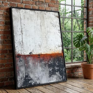 May include: Abstract art print in a black frame. The artwork features a textured surface with a white upper section, a rusty orange mid-section, and a dark gray lower section. The framed art is leaning against a brick wall.