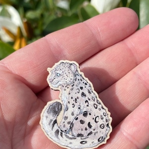 May include: A wooden snow leopard cut-out, showing a detailed illustration of the animal's spotted fur, blue eyes, and curled tail. The leopard is depicted in a seated position. The edges of the cut-out are smooth and the wood has a natural finish.