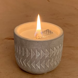 Könnte beinhalten: Ein grauer Beton-Kerzenhalter mit einer weißen Kerze und einem Metalldocht. Der Halter hat ein geometrisches Muster aus Dreiecken.