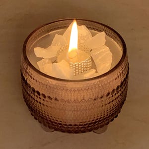 Könnte beinhalten: Eine brennende Kerze in einem dekorativen Glasbehälter. Die Kerze hat einen silberfarbenen Metallhalter und ist von weißen Wachsstücken umgeben. Der Behälter ist hellbraun mit einem strukturierten Muster und steht auf drei kleinen Füßen.
