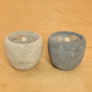 Könnte beinhalten: Zwei Beton-Kerzenhalter, einer grau und einer weiß, mit einer gesprenkelten Oberfläche. Jeder Halter hat eine kleine, runde, weiße Kerze darin.
