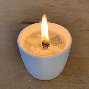 Könnte beinhalten: Ein weißer Kerzenhalter aus Keramik mit einer brennenden weißen Wachskerze. Die Kerze hat einen Docht aus Metall und ein Metallgitter oben.
