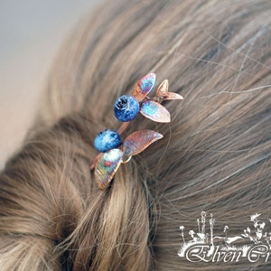 Puede incluir: Un peine para el cabello de cobre con bayas y hojas de vidrio azul. El peine está en el cabello de una mujer.