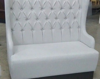 May include: A white, tufted-back bench with a high back and armrests. The bench has a dark brown base and is upholstered in a light-colored material. The back features a diamond-patterned button design.
