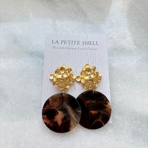 May include: Gold-toned floral stud earrings with large, marbled brown and tan circular pendants. The earrings are displayed on a white card with the text "LA PETITE SHELL" and the tagline "Naturally Charming Coastal Elegance."